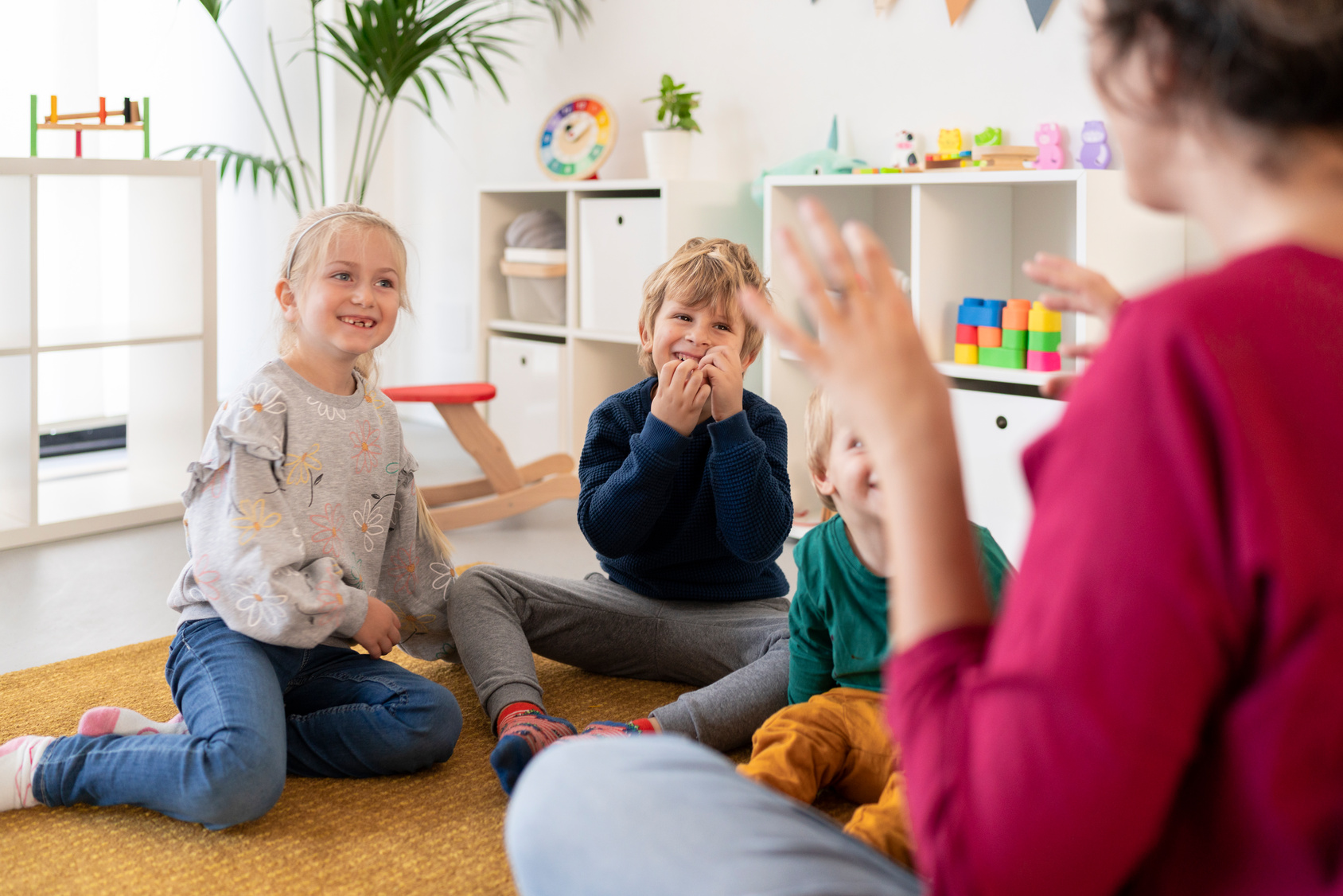 Smiling girl with arms raised sitting by boys while female teacher playing in preschool